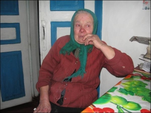 Марія Ментій сидить за столом у власній хаті в селі Соснівка Сосницького району Чернігівщини. Жінка ніколи не мала вдома телефону, майже не вміє по ньому говорити