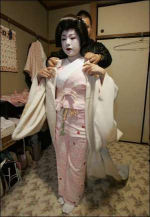 Асистент допомагає гейші Комомо одягти кімоно. Артистка має традиційний макіяж — вибілене обличчя, червоні щоки, підведені губи