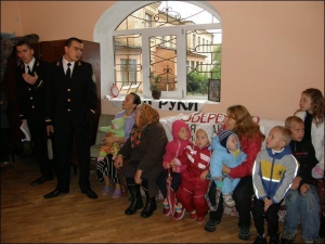 Многодетная мать Елена Лукашук с шестью своими детьми сидит на диване в доме Матусова. С правого края на подлокотник дивана присела жена Матусова, Наталия. С левой стороны на диване сидит мать Василия Матусова, а возле нее на стуле с младенцем на руках се