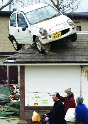 Троє японців стоять під будинком, на даху якого завис автомобіль. Біля споруди купи сміття, намиті хвилями цунамі. Місто Сендай чи не найбільше постраждало від стихії