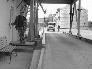 Охоронник елеваторного комплексу ”АгроБАР” у селі Міжлісся Барського району стоїть при вході до офісного приміщення. На задньому плані — завантажена зерном машина заїжджає на ваги 