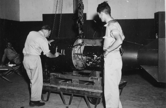 Командир А.Ф. Бирч (слева) нумерует бомбу под кодовым названием «Малыш» перед погрузкой ее на трейлер в здании Ассамблеи №1 перед конечной погрузкой бомбы на борт бомбардировщика B-29 Superfortress &quot;Enola Gay&quot; на базе 509-ой сводной группы на острове Тиниан у Марианских островов в 1945 году. Физик доктор Рамсей (справа) получит Нобелевскую премию в области физики в 1989 году. (U.S. National Archives)