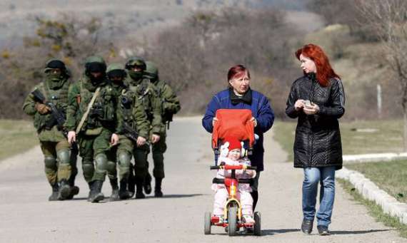 Жінки з дитиною у візку гуляють біля української військової бази в селі Перевальне під Сімферополем. Позаду – озброєні російські солдати у формі без розпізнавальних знаків. 14 березня 2014 року