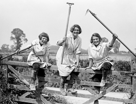 члени Жіночої аграрної армії посміхаються, піднімаючи свій інвентар в повітря і перелазячи через ворота, 1916