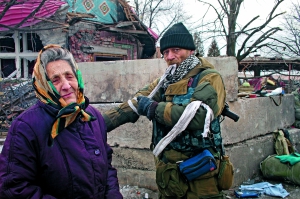 Сепаратист і літня жінка стоять біля зруйнованого будинку. Чоловік пов’язує собі на рукава біле ганчір’я. В такий спосіб бойовики розрізняють своїх