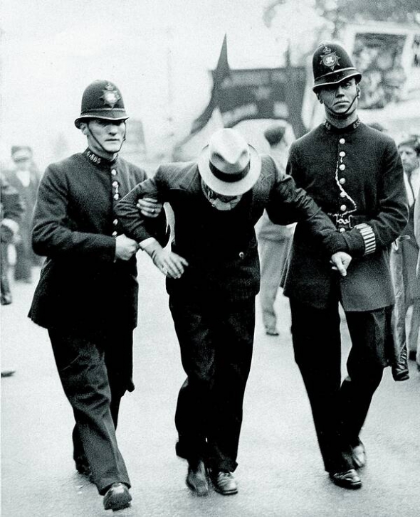 11 жовтня 1936 року поліцейські ведуть затриманого учасника маршу комуністів від пагорба Тавер-Гілл до парку Вікторія в Лондоні. Демонстранти вимагали заборонити у країні діяльність Британського союзу фашистів. Відбулися сутички між прихильниками двох політичних течій