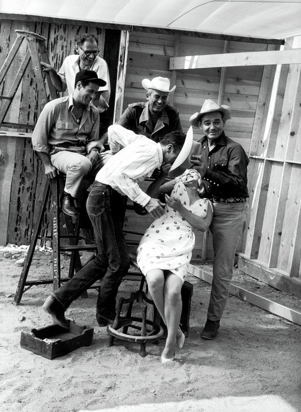 Мерилін Монро фотографується з колегами на знімальному майданчику в містечку Дейтон, штат Невада, у серпні 1960 року. Працювали над фільмом ”Неприкаяні”. Сценарій написав її чоловік Артур Міллер (стоїть на драбині). Їхні стосунки на той час розладналися і до виходу стрічки шлюб розпався. У картині знімався Кларк Ґейбл (у першому ряду праворуч). Казав, що білявка доведе його до нервового зриву. За 11 днів після зйомок помер