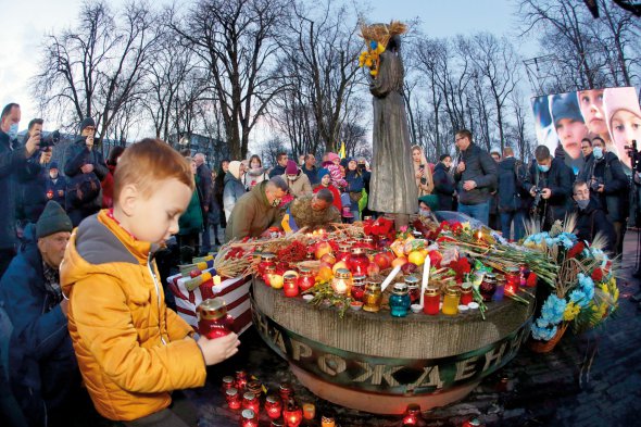 Люди стоять біля меморіалу пам’яті жертв Голодомору в Києві увечері 27 листопада. До пам’ятника ”Гірка пам’ять дитинства” несуть свічки, хліб, зерно, калину та яблука