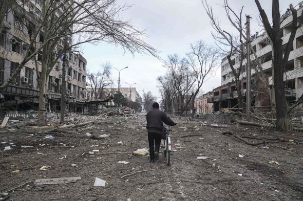 Чоловік йде з велосипедом по вулиці Маріуполя. Навкруги зруйновані житлові будинки та повалені дерева через авіаудари окупантів. Місто обстрілюють та бомбардують майже щодня