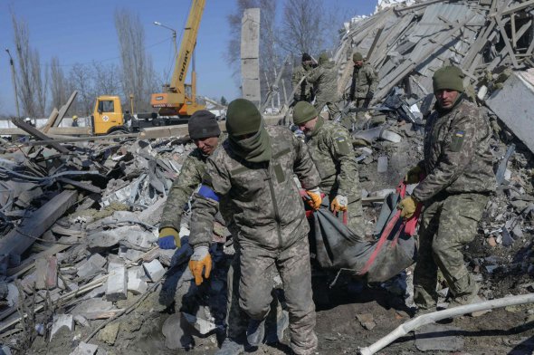 Солдати виносять мертвого бійця з-під уламків казарми в Миколаєві, 19 березня 2022 року. Напередодні будівлю зруйнували російські ракети типу ”Калібр”. Потрапили в місце постійного дислокування 79-ї окремої десантно-штурмової бригади Збройних сил України. Про атаку не встигли попередити, бо вона відбулася з близької відстані — з Херсонщини. Загинули 50 українських бійців.