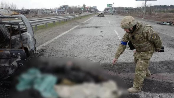 Украинский воин указывает на обгоревшие человеческие останки возле сгоревшего автомобиля.