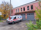 Сотрудник отделения экстренной медицинской помощи Изюмской городской больницы моет автомобиль возле поврежденного обстрелами здания