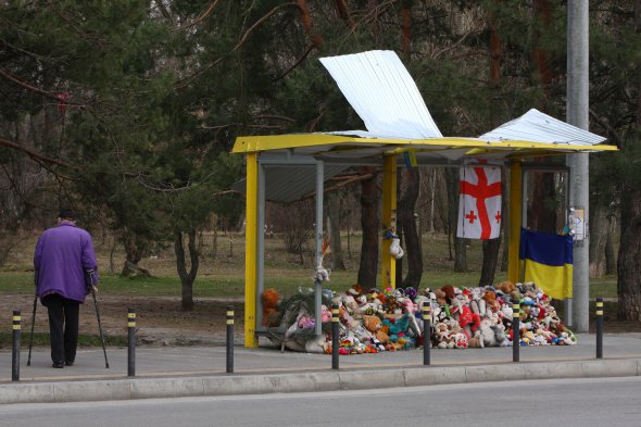 Зупинка навпроти зруйнованого будинку. Люди досі приносять сюди іграшки й квіти, щоб вшанувати памʼять загиблих