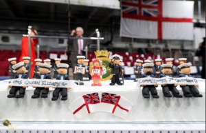 Специально для церемонии изготовили торт, символизирующий авианосец. В центре - капитан корабля Джерри Крид, матрос, который держит табличку с надписью HMS и британская королева, которая держит табличку с надписью "Я"