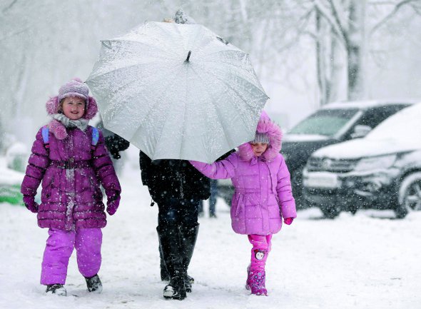 Жінка з двома дівчатами йде Києвом під час хуртовини 18 грудня. Закривається від снігу парасолькою. Сильні опади у столиці почалися в понеділок зранку. Утворилися кілометрові затори, погано ходив транспорт. Розчищати дороги виїхали майже 450 одиниць комунальної техніки