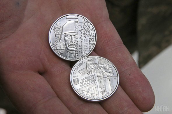 Монета имеет композицию с изображением защитника аэропорта на фоне полуразрушенной башни, над которой развевается украинский флаг