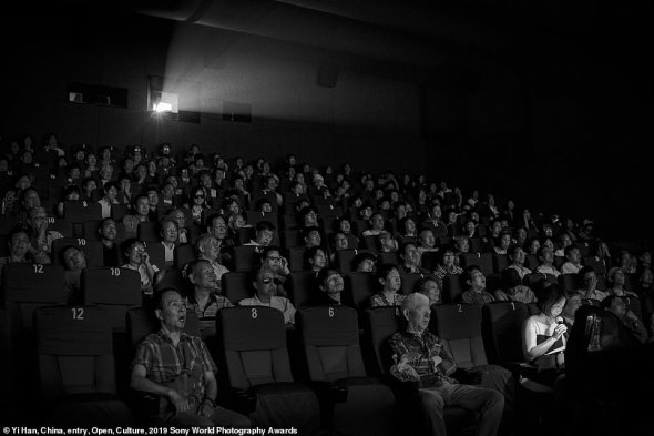 На цьому фото зображений момент, коли група сліпих людей і тих, які погано бачать  "дивляться" фільм в театрі Шанхаю. У Китаї 18 мільйонів сліпих людей - це найбільше число сліпих серед країн світу. Волонтерські організації придумують способи, як зробити кіно більш доступним для таких людей. Вони придумали робити живий звуковий опис. Оповідач сидить в кімнаті перегляду і розповідає, що відбувається на екрані. Фотографію зробили в травні 2018 року в Історичному театрі в Шанхаї