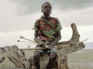 14-річний Ману, одягнений в сорочку з кольорами ямайського прапора, тримає чотири стріли і цибулю, сидячи на Мертвому дереві в Танзанії