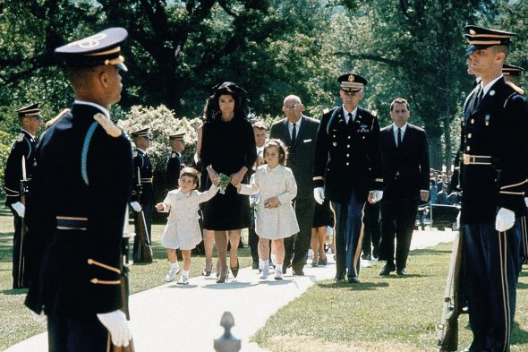 Вдова президента США Джона Фіцджеральда Кеннеді 34-річна Жаклін приїхала 29 травня 1964-го на Арлінгтонське національне кладовище в передмісті Вашингтона. Із нею діти – 4-річний Джон та 7-річна Кароліна. Того дня її чоловіку виповнилося б 47 років. Його вбив зі снайперської гвинтівки відставний морський піхотинець Лі Харві Освальд. Це сталося в листопаді 1963-го в Далласі. Через чотири роки Жаклін Кеннеді вийшла заміж за грецького мільярдера Аристотеля Онассіса. Працювала редактором різних видань. Син став адвокатом, а донька – письменницею. 1975-го Жаклін овдовіла вдруге. Жила сама. Померла 1994-го від раку