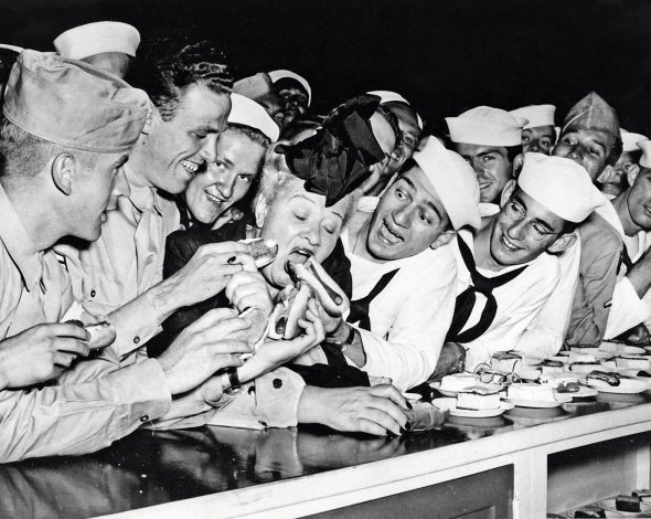 Моряки годують хот-догами американську співачку 57-річну Софі Такер у Чикаго 2 липня 1944-го. Беруть участь у відкритті клубу Об’єднаних організацій обслуговування збройних сил. Ця спільнота збирала благодійні внески та організовувала дозвілля військових