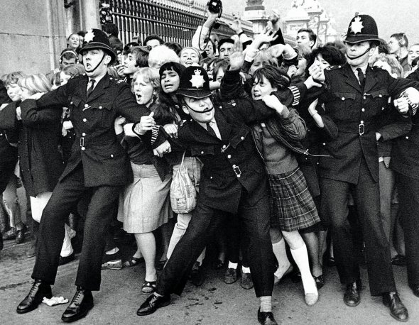 Фанатки британської рок-групи The Beatles намагаються прорвати поліцейський кордон біля Букінгемського палацу в Лондоні 29 липня 1965-го. Того дня в кінотеатрі ”Павільйон” на площі Пікаділлі відбулася прем’єра фільму Help! У ньому знялися музиканти гурту. Тисячі фанатів вишикувалися вулицями, очікуючи кумирів, які приїхали в чорному Roll’s-Royce. У залі була королева Великої Британії 39-річна Єлизавета II. Після того вона нагородила музикантів орденами